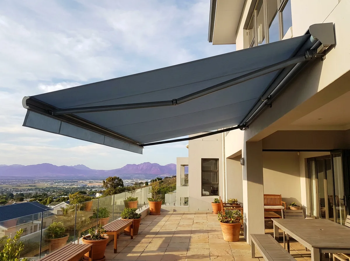 A folding-arm awning extended on folding metal arms over a stone entertaining terrace at an elevated Welgemoed home, throwing overhead shade, with a breezy outlook over the Cape Flats and distant mountains beyond
